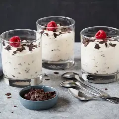 Drei Gläser mit cremigem Dessert, garniert mit Himbeeren und Schokoladenstückchen. Ein Schälchen mit Schokostückchen und Löffel daneben. Ein leckeres Herrencreme Rezept zum Nachmachen.
