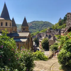 Un pueblo pintoresco con una iglesia románica y tejados de pizarra, ideal para rutas por Francia. Flores rosas y verdes exuberantes en primer plano.