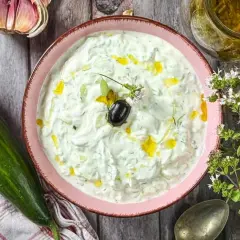 Schüssel mit cremigem griechischem Tzatziki, garniert mit Olive und Öl. Daneben Gurke, Knoblauch und frische Kräuter.