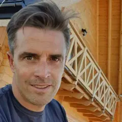 Staszek Karpiel-Bułecka pozuje na tle drewnianego domu z balustradą.