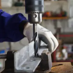 Ręka w rękawicy obsługuje wiertarkę stołową, wiercąc metalowy profil. Widać wiertło i wióry.