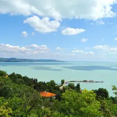 Widok na turkusowe jezioro Balaton z zielonymi wzgórzami i domami. Na wodzie widać molo i żaglówki.