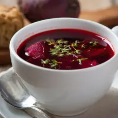 Aromaticzny barszcz czerwony domowy, z kawałkami buraka i ziołami, podany w białej filiżance z łyżeczką.