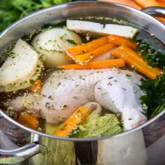 Pentola con brodo di pollo ricetta della nonna: pollo, carote, sedano, cipolla e prezzemolo in cottura su un tavolo di legno.