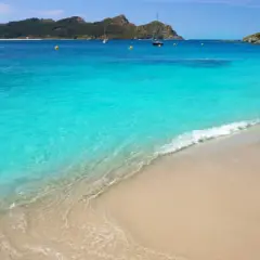 Aguas turquesas y arena dorada en unas playas cristalinas España, con veleros navegando al fondo y montañas verdes.