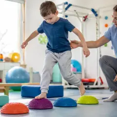Chłopiec ćwiczy równowagę na poduszkach sensorycznych, a fizjoterapeutka asekuruje go. To część terapii, która może przypominać grę w boccia.