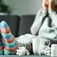 Osoba w kolorowych skarpetkach chora, otulona kocem, z chusteczkami i lekami. Objawy alergii dają się we znaki.