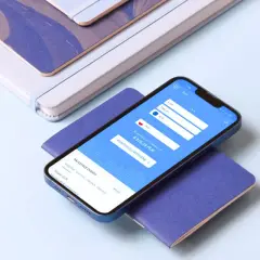 Smartfon z aplikacją do wymiany walut HKD na PLN, obok pióro i notatniki.