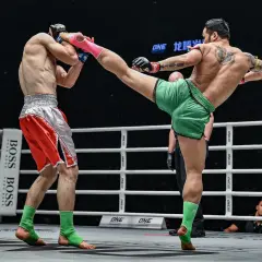 A Muay Thai fighter in green shorts lands a powerful kick to his opponent's head. The opponent, in red shorts, braces for impact, his foot nearly touching the canvas.