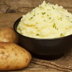 Un cremoso puré de patatas Thermomix, adornado con perejil fresco, junto a patatas crudas sobre una mesa de madera.