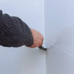 Ręka w swetrze usuwa stary silikon z białych płytek za pomocą narzędzia. Ocet może pomóc w usuwaniu silikonu.
