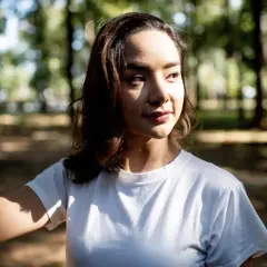 A young woman in a white t-shirt stands in a park, her face illuminated by dappled sunlight, showcasing the dramatic contrast of soft lighting vs hard lighting.