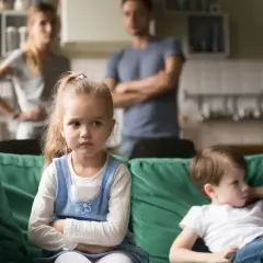 Smutne dzieci na kanapie, rodzice w tle. Widoczna zazdrość u dzieci w wieku szkolnym, gdy czują się pominięte.