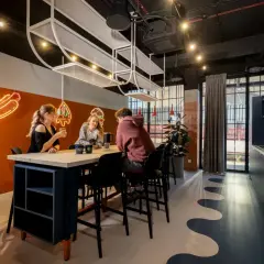 Studenci przy stole, rozmawiają i piją napoje. W tle neonowe hot dogi i pizza. To miejsce sprzyja nauce i relaksowi.