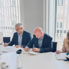 Cztery osoby przy stole podpisują dokumenty. W tle okna z żaluzjami. To spotkanie dotyczące triglav ubezpieczenia.