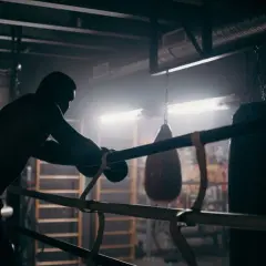 A boxer leans on the ropes in a dimly lit gym, contemplating how to get into boxing matches. Punching bags hang in the background.