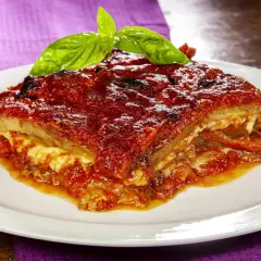 Una fetta di parmigiana di melanzane ricetta originale, ricca e succulenta, guarnita con basilico fresco, su un piatto bianco.