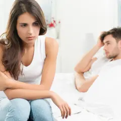 Smutna kobieta siedzi na łóżku, zmartwiona. Jej partner leży obok, z ręką na czole. Czy to uczulenie na spermę powoduje ich problemy?