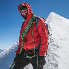 Uśmiechnięty alpinista w czerwonej kurtce na szczycie góry, z liną na ramieniu, na tle majestatycznego **Mount Blanc**.