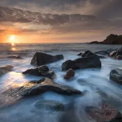 Available light photos capture a serene sunset over a rocky coastline. Water flows gently around dark, wet stones, reflecting the warm hues of the sky.