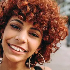 Sonrisa radiante de una joven con un vibrante castaño rojizo pelo rizado, luciendo grandes aros plateados.