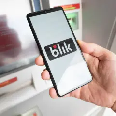 Dłoń trzyma smartfon z logo Blik przed bankomatem. Uważaj na oszustów, którzy mogą próbować wyłudzić dane.