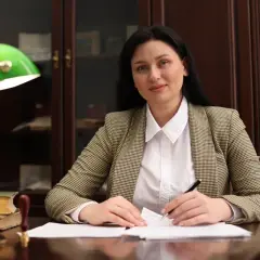 Kobieta w marynarce siedzi przy biurku, pisząc piórem na papierze, obok lampka i książki.