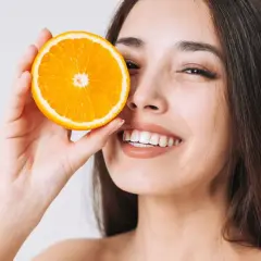 Lächelnde Frau hält Orangenscheibe vor ihr Auge. Vitamin C kann bei Rosacea helfen, die Haut zu beruhigen.