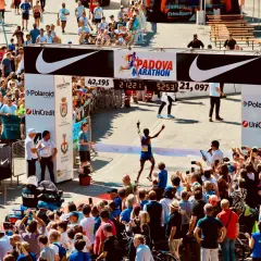 Meta-brama z napisem "Padova Marathon" i biegnącym zawodnikiem, tłum widzów, zegar z czasem.