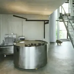 Interno in stile brutalista con cucina circolare in metallo, scale a vista e pareti in cemento grezzo.
