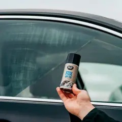 Dłoń trzyma puszkę K2 FOX ANTI-FOG, która pomoże Ci jak odparować szyby w aucie. Widoczne smugi na szybie sugerują problem z zaparowaniem.