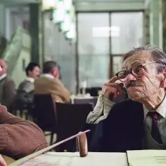 Dwóch mężczyzn rozmawia przy stole. Jeden z nich, starszy, w okularach, gestykuluje. Scena z jednego z filmów Krzysztofa Krauzego.