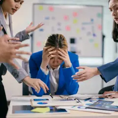 Giovane donna d'affari stressata, con le mani sulla testa, mentre colleghi la circondano con gesti agitati. Lo stress lavoro correlato è un rischio da valutare.