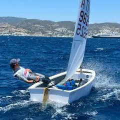 Giovane velista inclinato per bilanciare la piccola imbarcazione, che solca le onde blu. Sullo sfondo, una costa con case e una nave da crociera. La traccia delle barche è visibile nell'acqua.