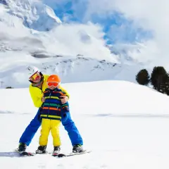 Un adulto insegna a sciare a un bambino di 2 anni su una pista innevata, con montagne sullo sfondo. Un perfetto weekend con bambini 2 anni!