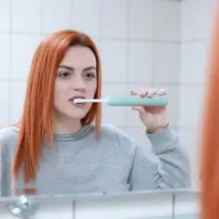 Ruda kobieta w szarej bluzie myje zęby szczoteczką soniczną przed lustrem. Pokazuje, jak myć zęby szczoteczką soniczną.