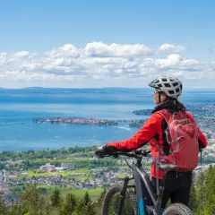 A woman in a red jacket and helmet pauses her **Lake Constance bike tour** to admire the expansive blue lake and distant towns under a cloudy sky.