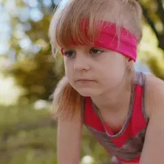 Zmęczona dziewczynka w sportowym stroju, z opaską na głowie, pochyla się, widać, że dziecko szybko się męczy.