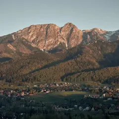 Widok z **Antałówki punkt widokowy** na Tatry. Słońce oświetla skaliste szczyty i lasy, a w dole widać zabudowania.