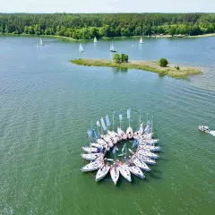 Żaglówki tworzą okrąg na zielonych jeziorach mazurskich, otoczone lasem.