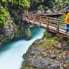 Ein Wanderer auf einer Holzbrücke über einem türkisfarbenen Fluss in einer Schlucht, ähnlich dem Bleder See.