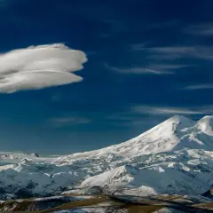 Pokryty śniegiem najwyższy szczyt Kaukazu, Elbrus, góruje nad ośnieżonymi pasmami górskimi pod błękitnym niebem z charakterystyczną chmurą.
