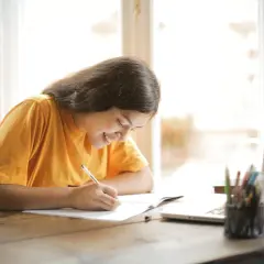 Uśmiechnięta dziewczyna pisze w zeszycie, przygotowując się do zadanie 1 matura ustna angielski. Obok laptop i przybory do pisania.