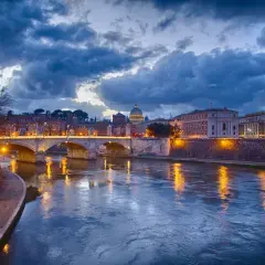 Il Tevere riflette le luci di Roma al tramonto, con la cupola di San Pietro sullo sfondo. La **Trastevere nightlife** è già nell'aria, promessa di serate vivaci.