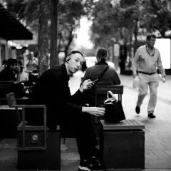 A man with earbuds looks at his phone while sitting on a bench. This 35mm vs 50mm street photography captures a moment of urban life.