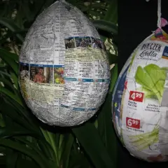Jak zrobic jajko z gipsu w balonie? Dwa papierowe jajka, jedno z gazet, drugie z reklam, wiszą na tle zielonych liści.