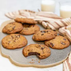 Nybakade chocolate chip cookies recept på en grå platta, med en bit tagen ur en cookie. Två glas mjölk och en randig handduk i bakgrunden.