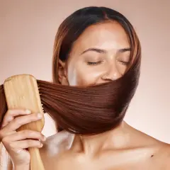 Frauen mit geschlossenen Augen bürsten ihr glänzendes, braunes Haar mit einer Holzbürste. Ein Moment der Ruhe und des **Haare pflegen**.
