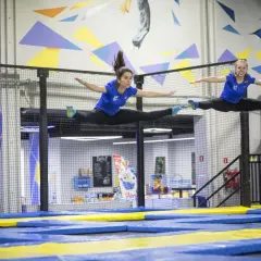 Dwie kobiety w niebieskich koszulkach wykonują szpagat w powietrzu na trampolinach.