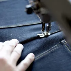 A sewing machine stitches a precise jean seam on dark denim, with a hand guiding the fabric.